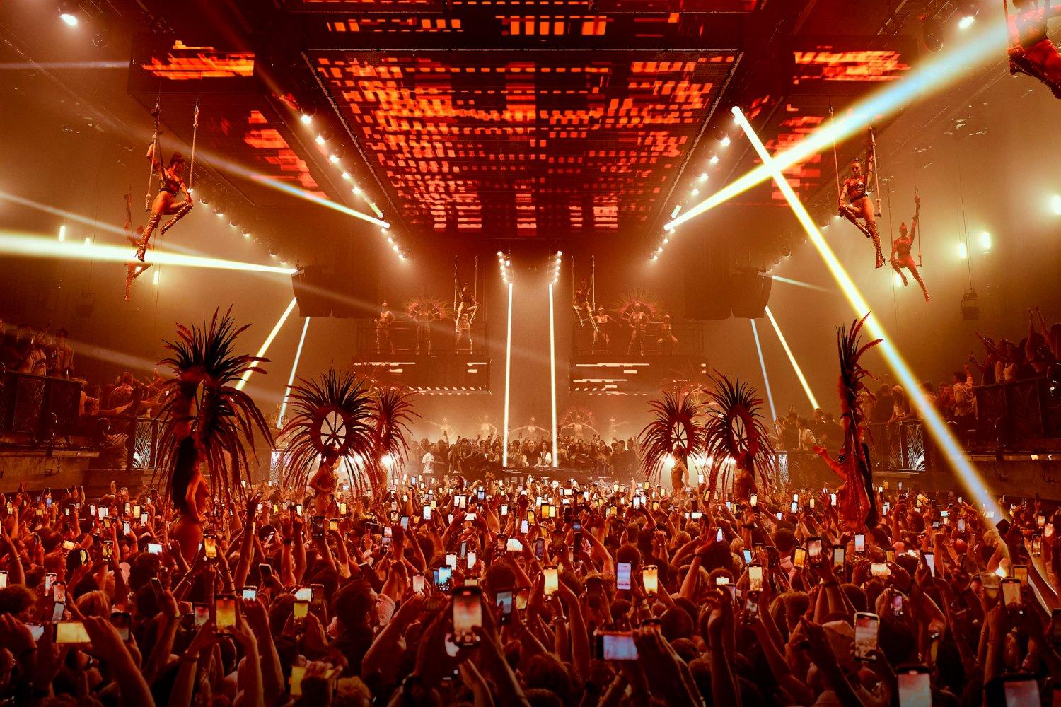 Vibrant Ibiza club scene with a large crowd, aerial performers, and red stage lighting.