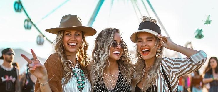 Three smiling women in stylish festival outfits, including hats and layered jewelry, with a Ferris wheel in the background.