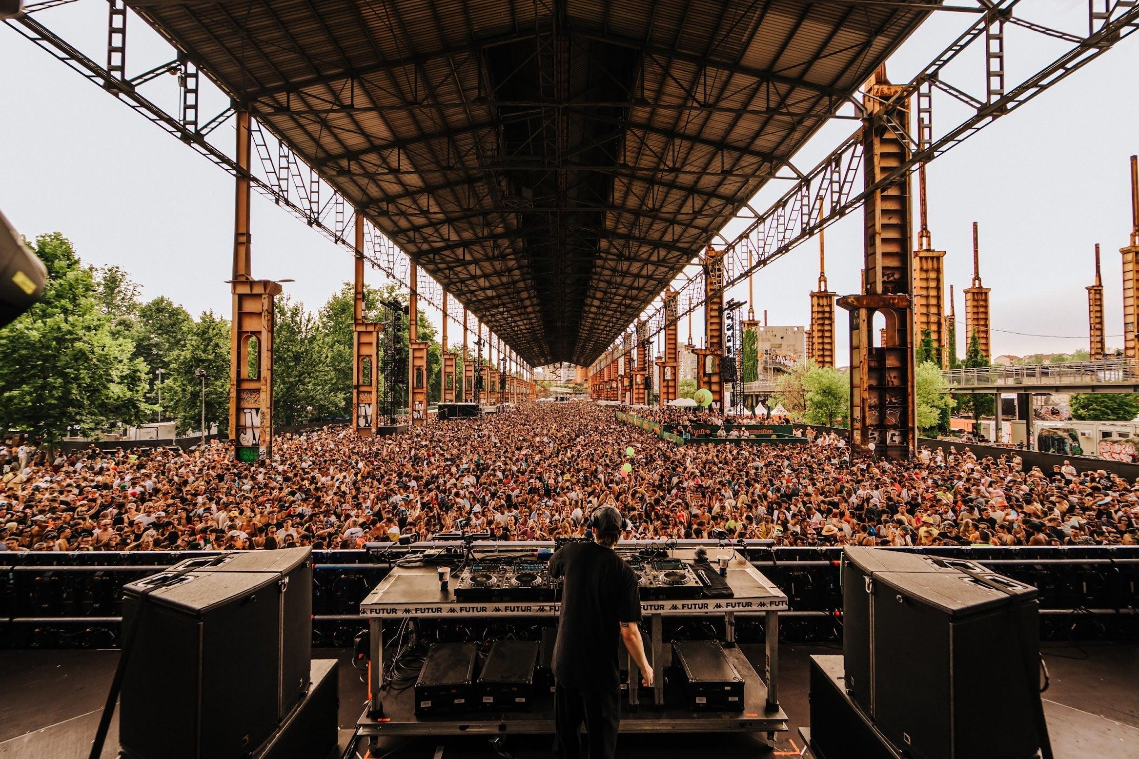 DJ on stage facing a massive crowd under a long industrial structure at Kappa FuturFestival.