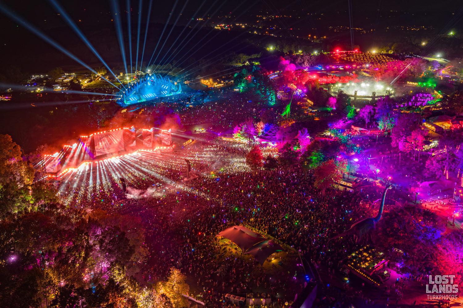Aerial view of a massive outdoor music festival at night, with vibrant laser lights illuminating large crowds and stages.