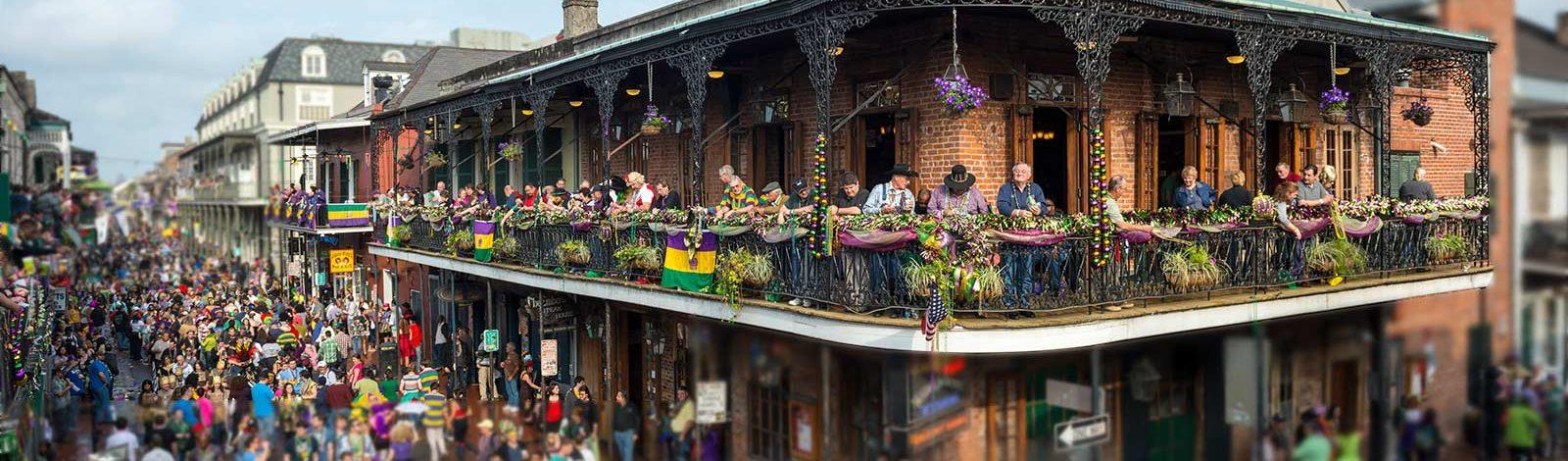 Crowd gathered for Mardi Gras in New Orleans, with people on a wrought-iron balcony overlooking the street.