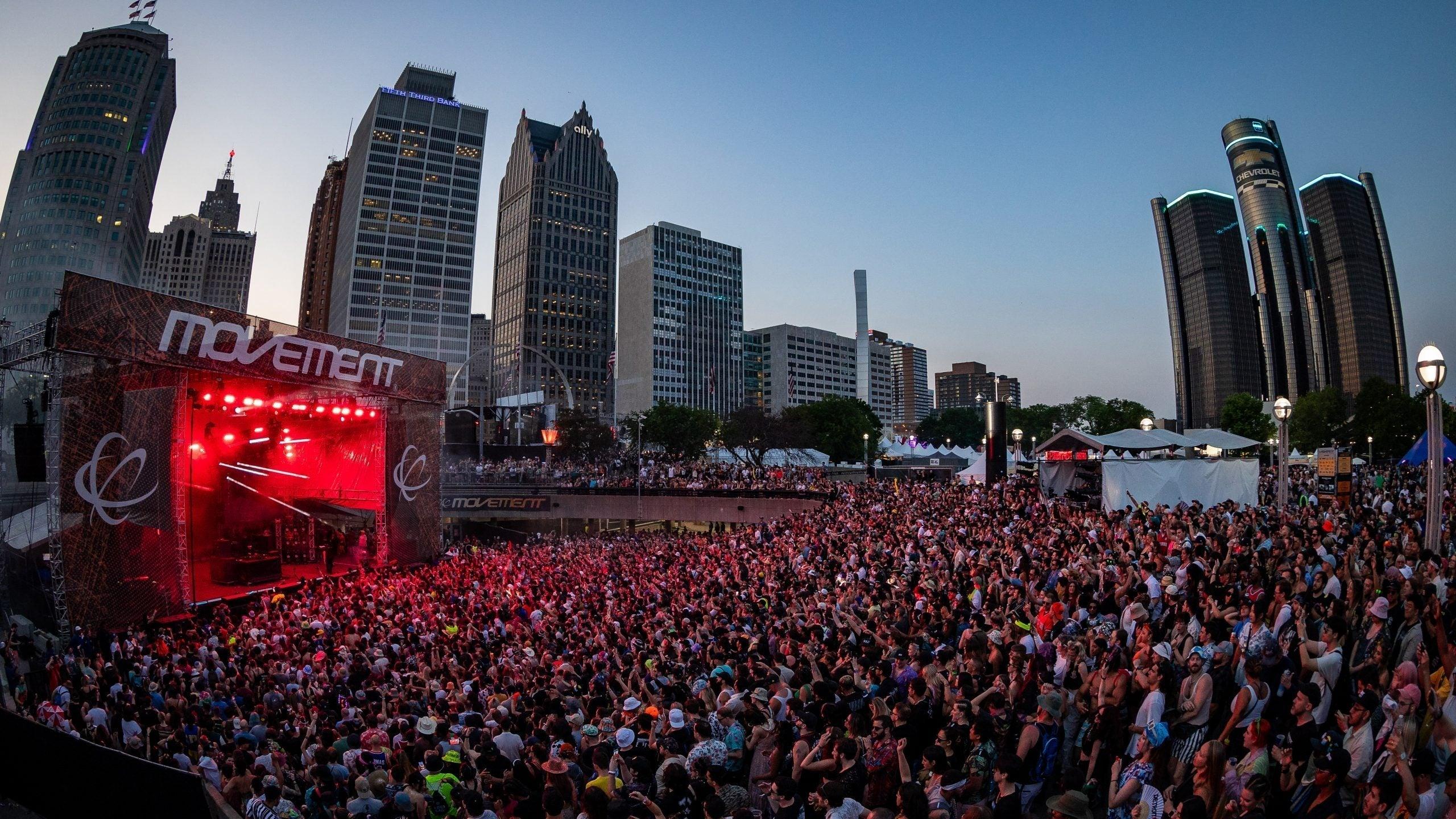 Crowd at Movement Detroit festival in Hart Plaza with city skyline at dusk, red stage lights.