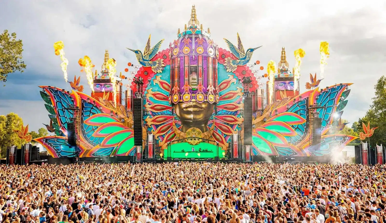 Vibrant Mysteryland main stage with a large golden face, colorful wings, and pyrotechnics over a massive crowd.