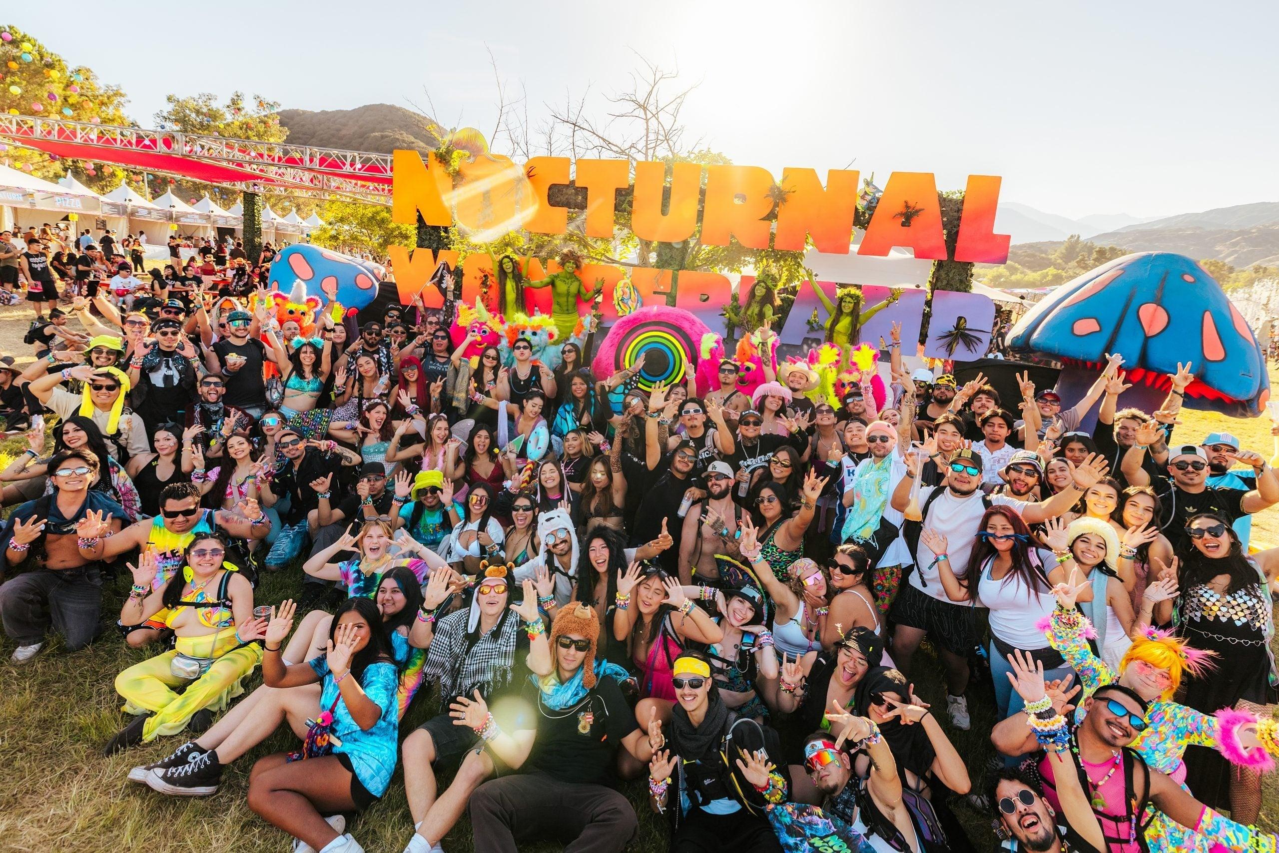 Large crowd of festival-goers posing in front of "Nocturnal Wonderland" sign and colorful mushroom art.
