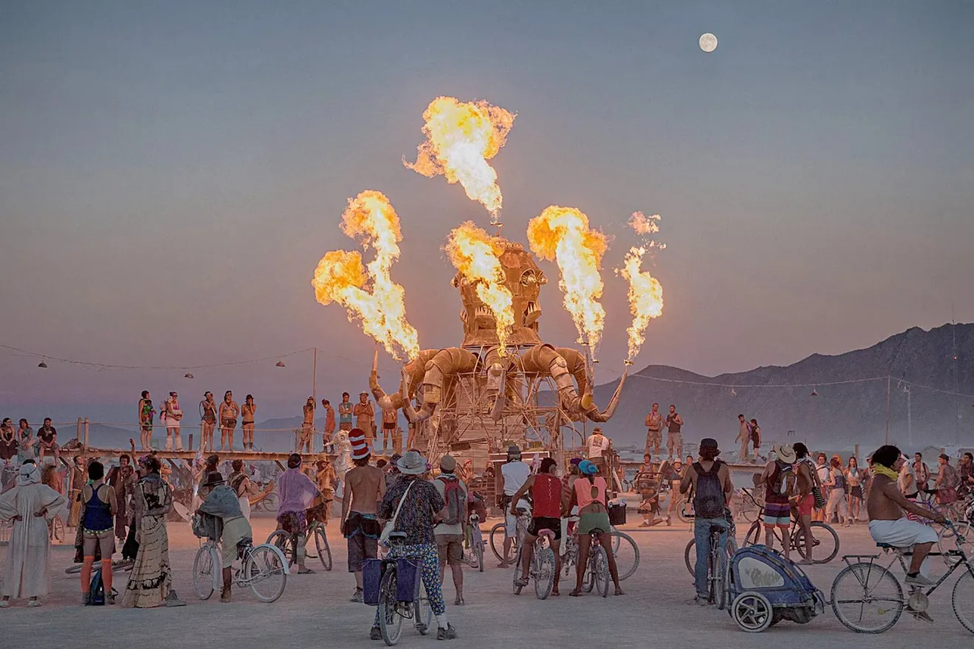 Festival-goers gather around a large, burning octopus sculpture under a full moon at dusk.