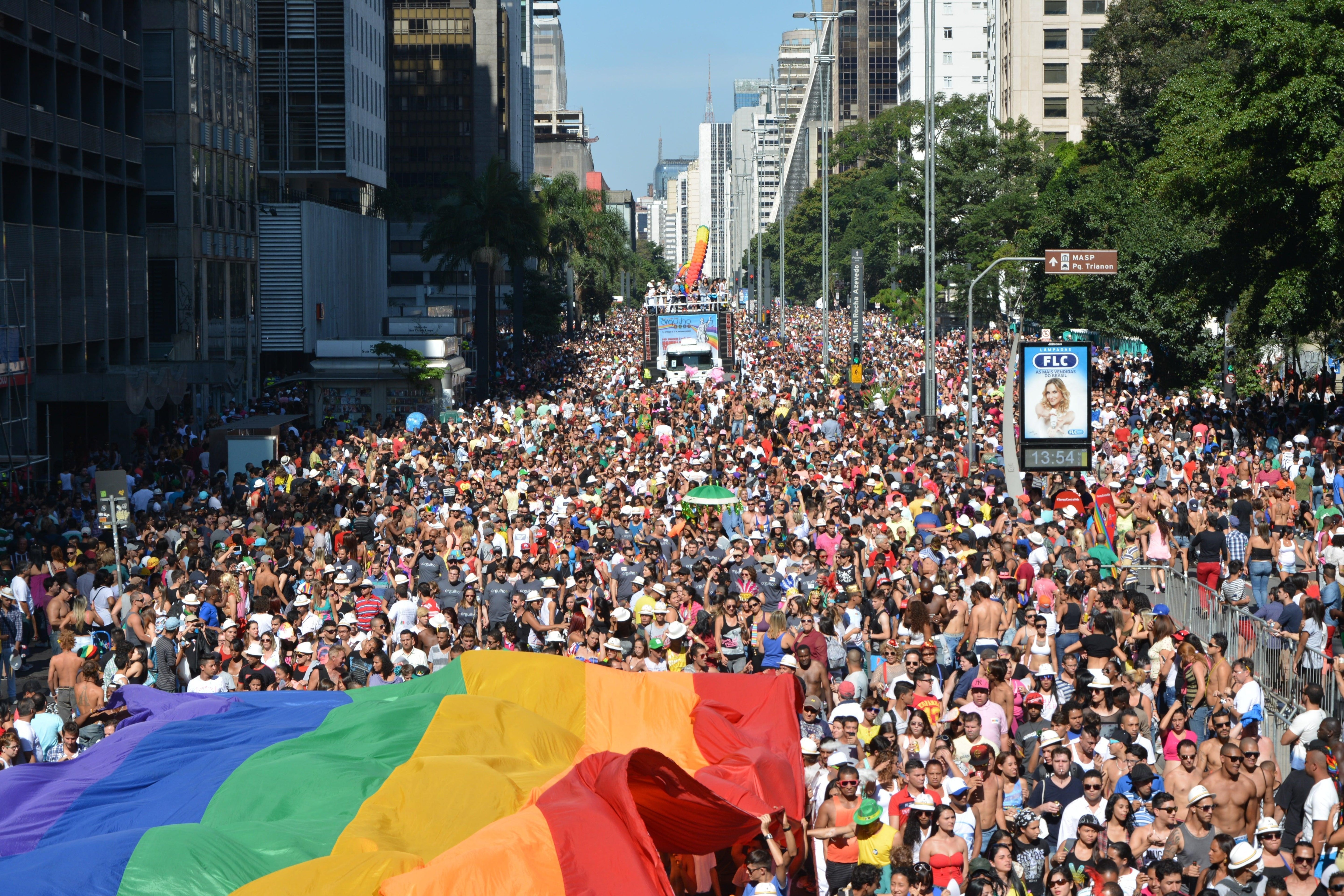 Massive crowd at São Paulo Pride, with a large rainbow flag in the foreground and parade floats.