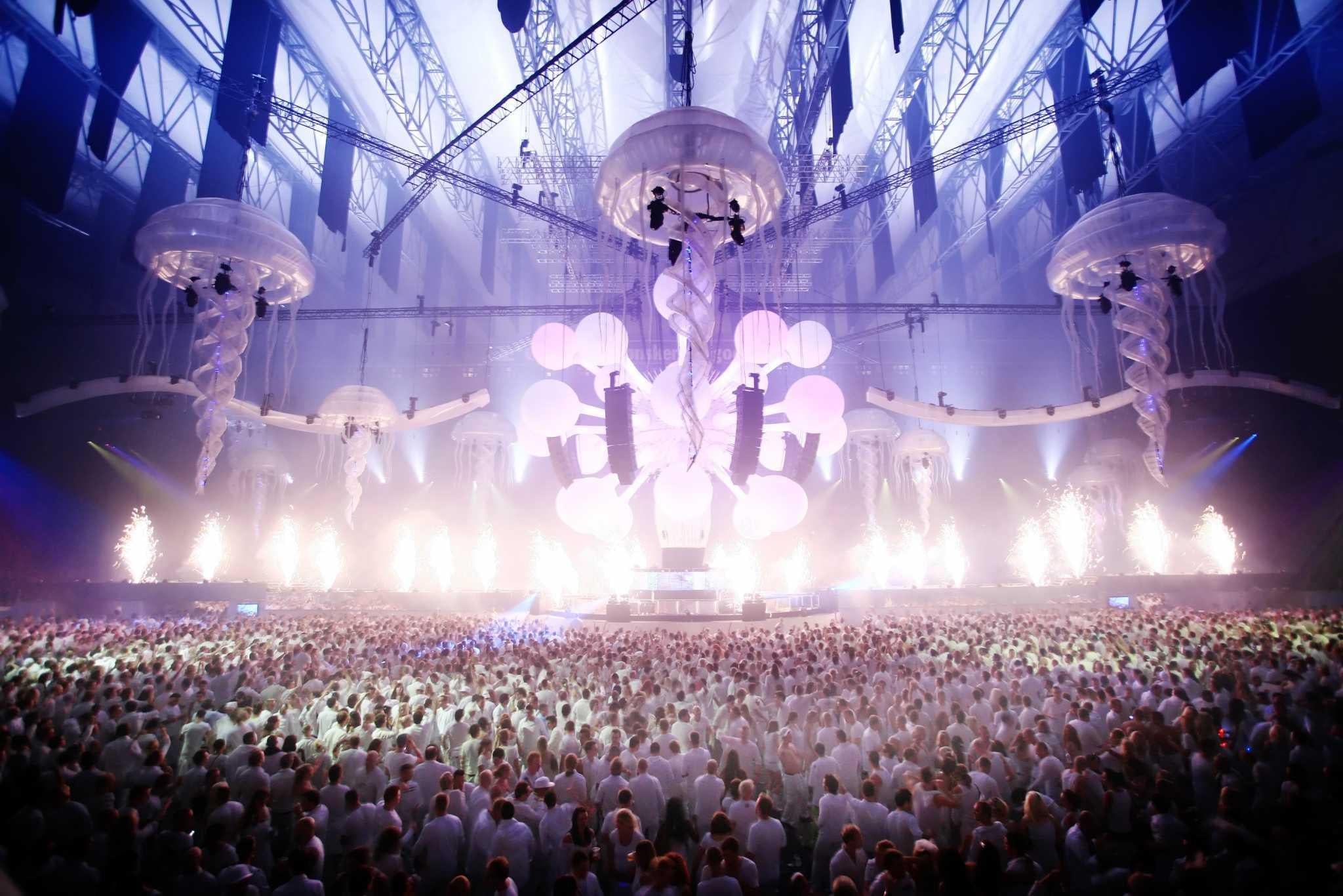 Vast crowd in white attire at Sensation Amsterdam, under glowing jellyfish decor and bright stage lights.