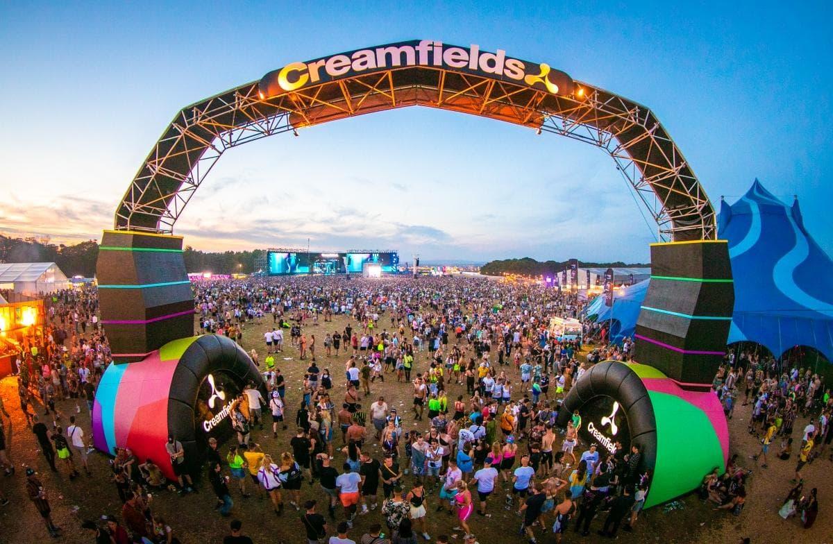 Creamfields festival entrance arch shaped like headphones, glowing at dusk over a large crowd.