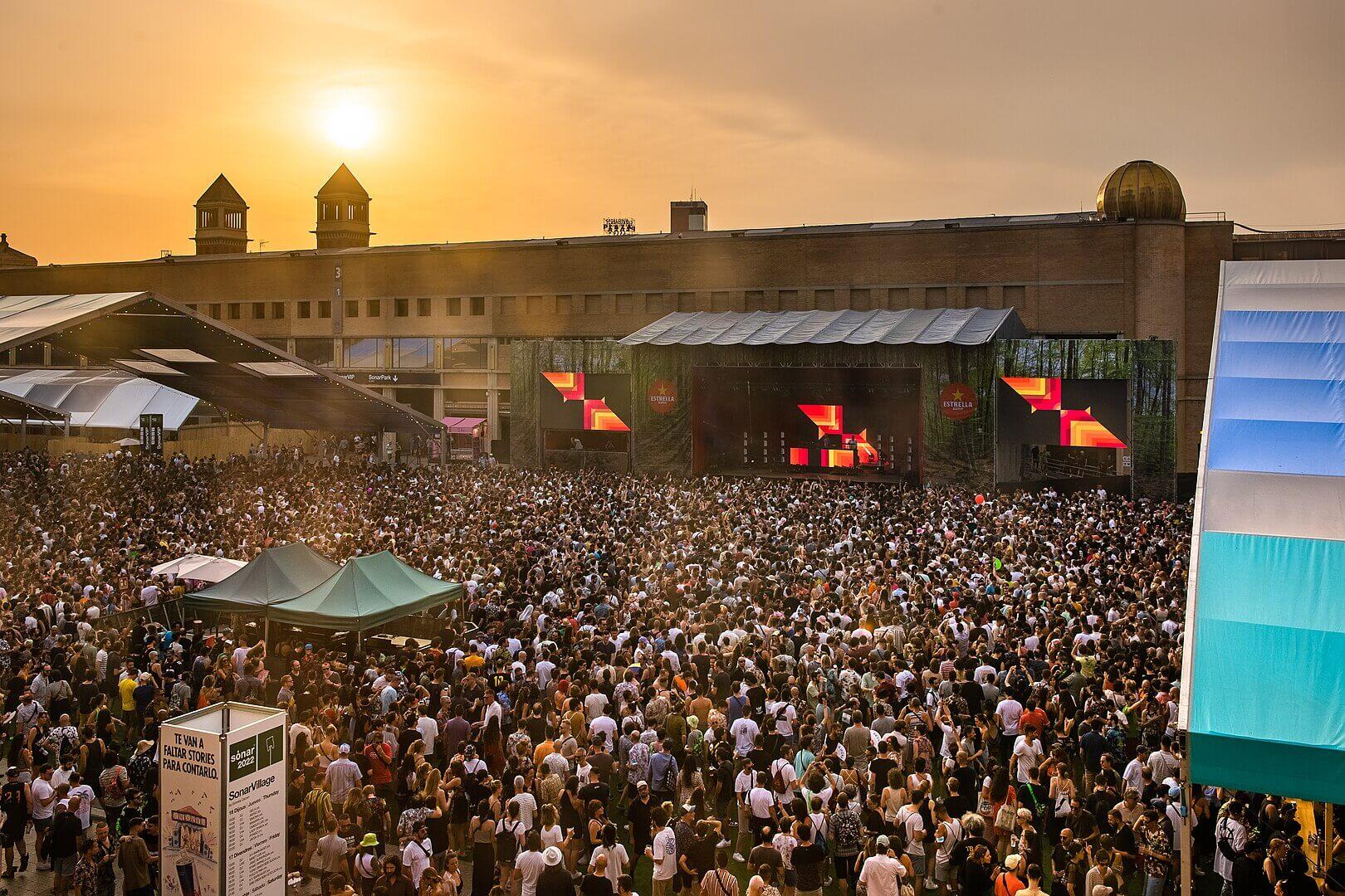 Crowd at an outdoor electronic music festival with a large stage and sunset glow.