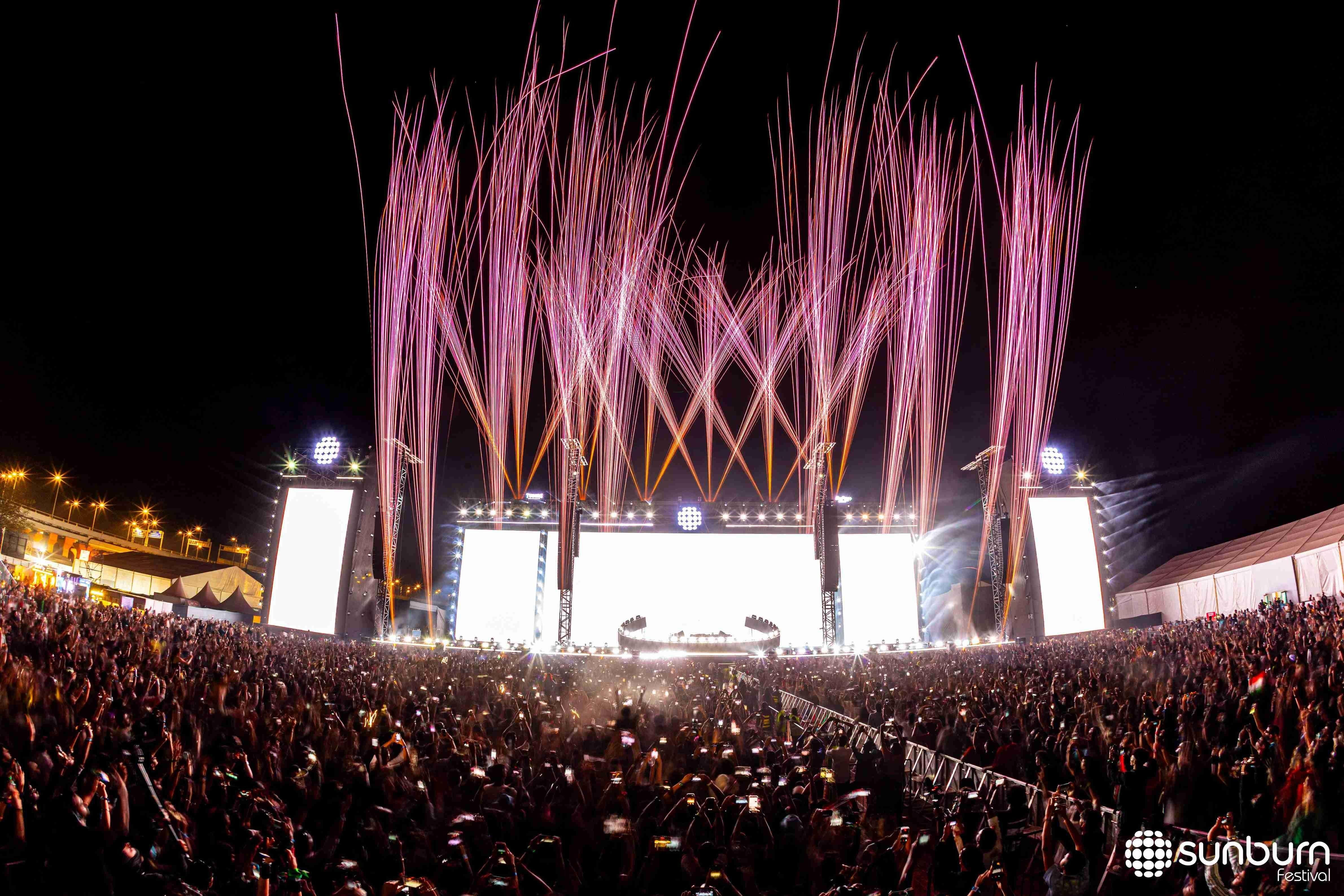 Massive outdoor EDM festival stage with bright screens, pink fireworks, and a huge crowd at night.