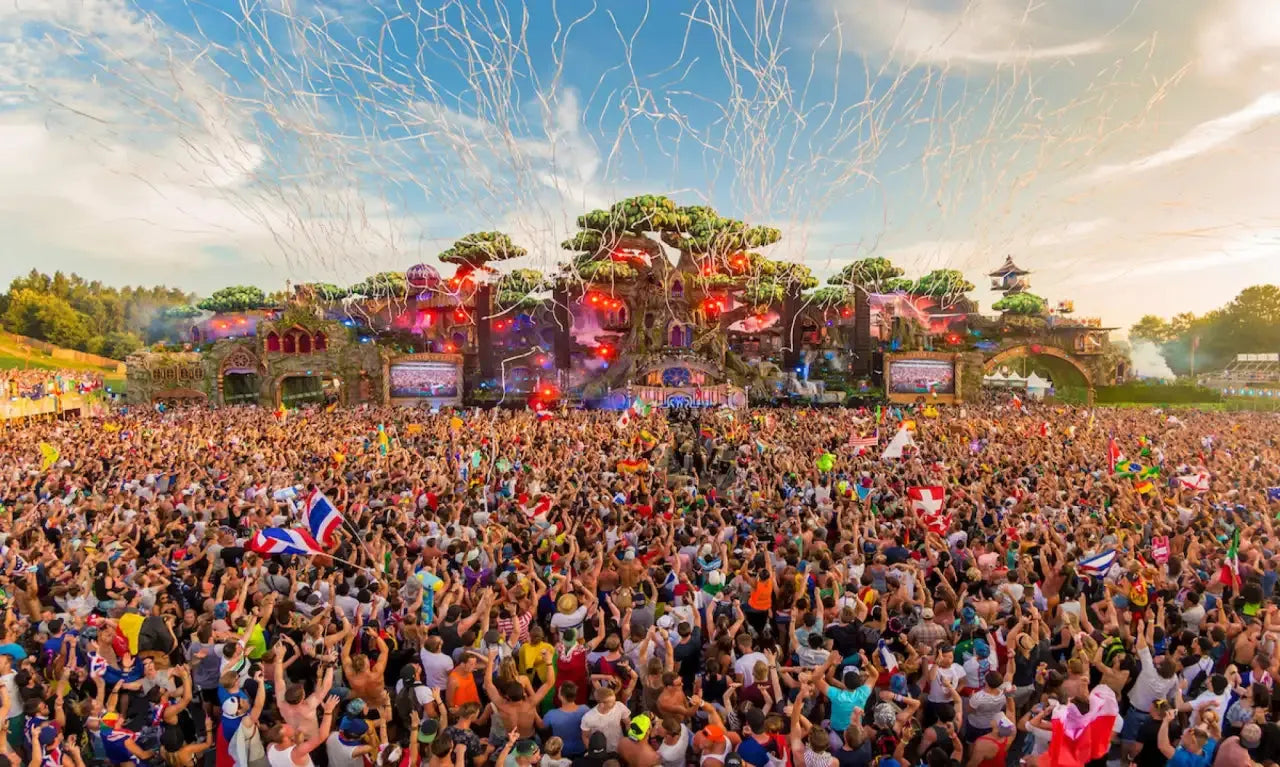 Massive EDM festival crowd with confetti, a large stage, and fantasy-themed decorations.