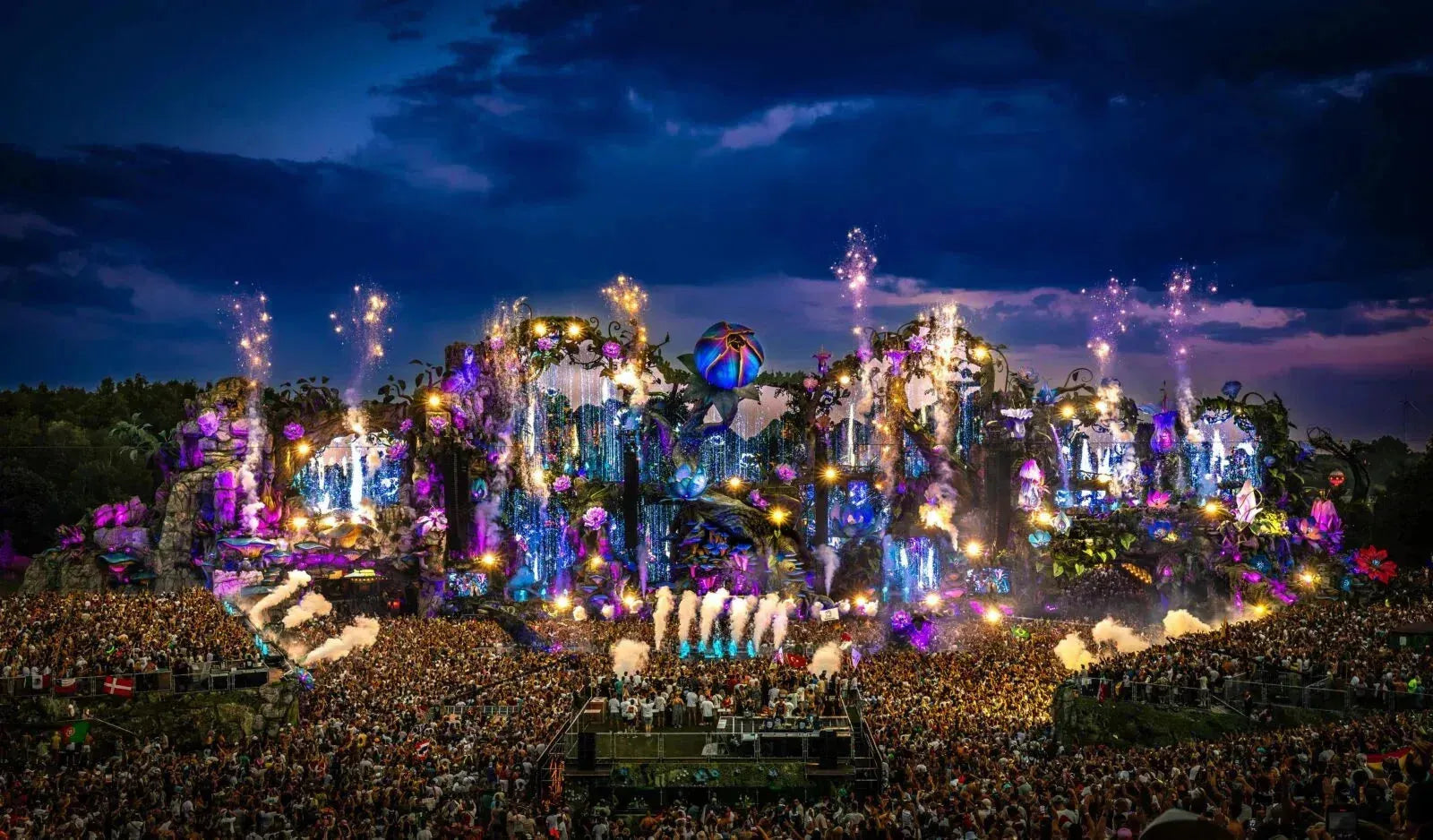 Tomorrowland main stage at dusk, vibrant with purple and blue lights, fireworks, and a massive crowd.