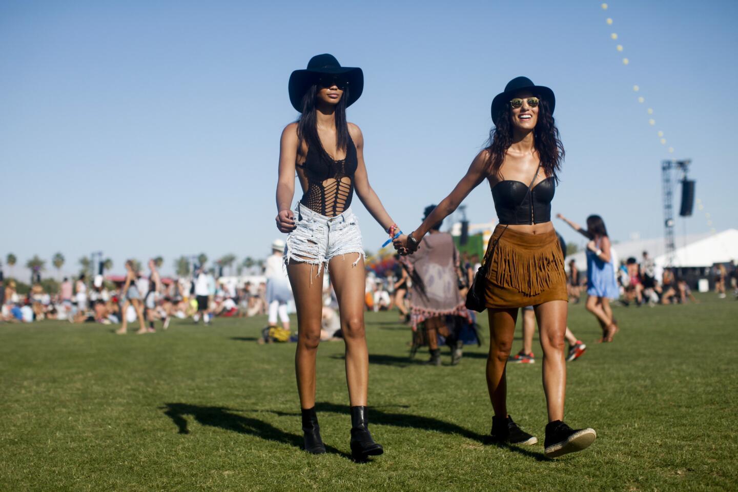 Two women in stylish festival outfits: black hats, distressed denim shorts, and a fringe skirt.