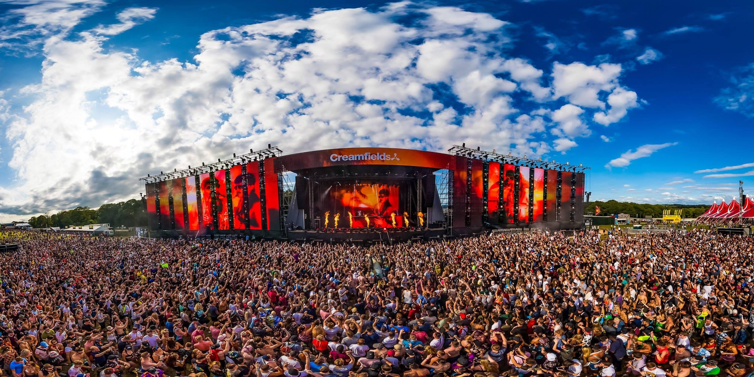 Vast crowd at Creamfields festival, hands raised, facing a large stage with vibrant red screens and performers.