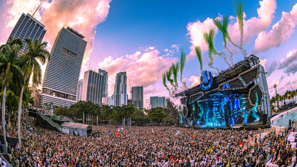 Massive crowd at Ultra Music Festival with a vibrant main stage, green pyro, and Miami skyline at sunset.