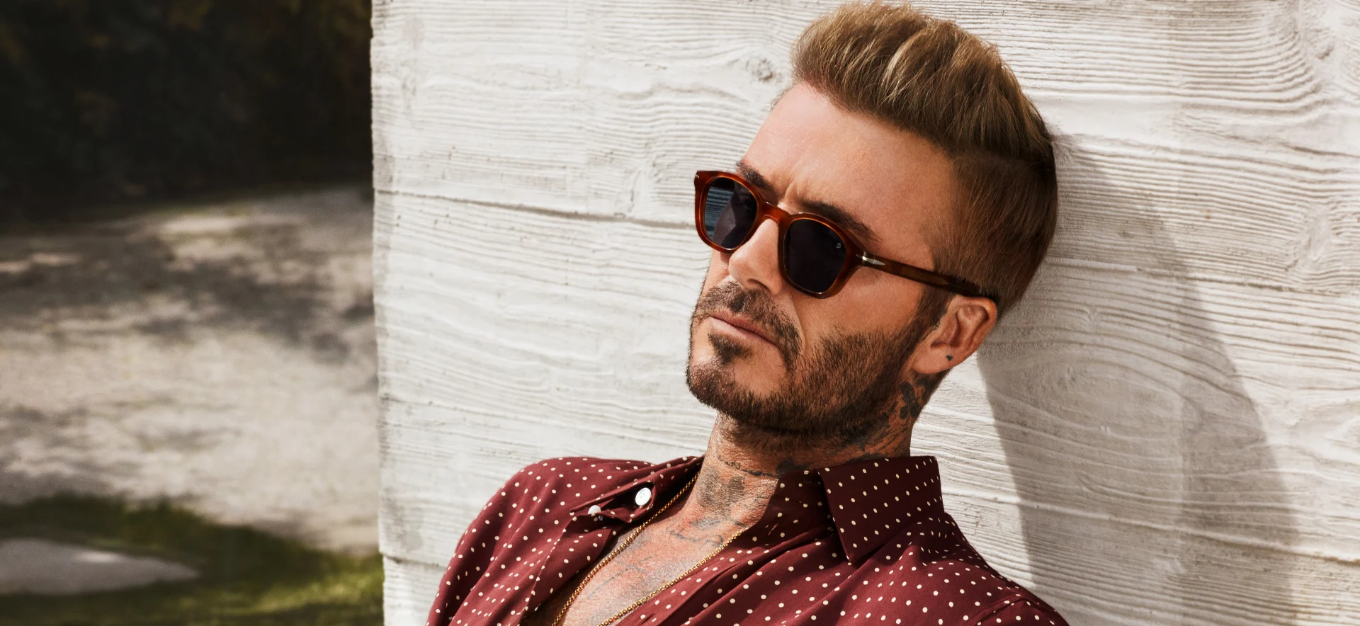 David Beckham in stylish brown tortoiseshell sunglasses, a red polka dot shirt, leaning on a textured wall.