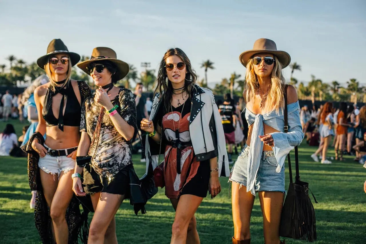 Four women in stylish festival outfits, including hats, sunglasses, and layered clothing, walking on grass.