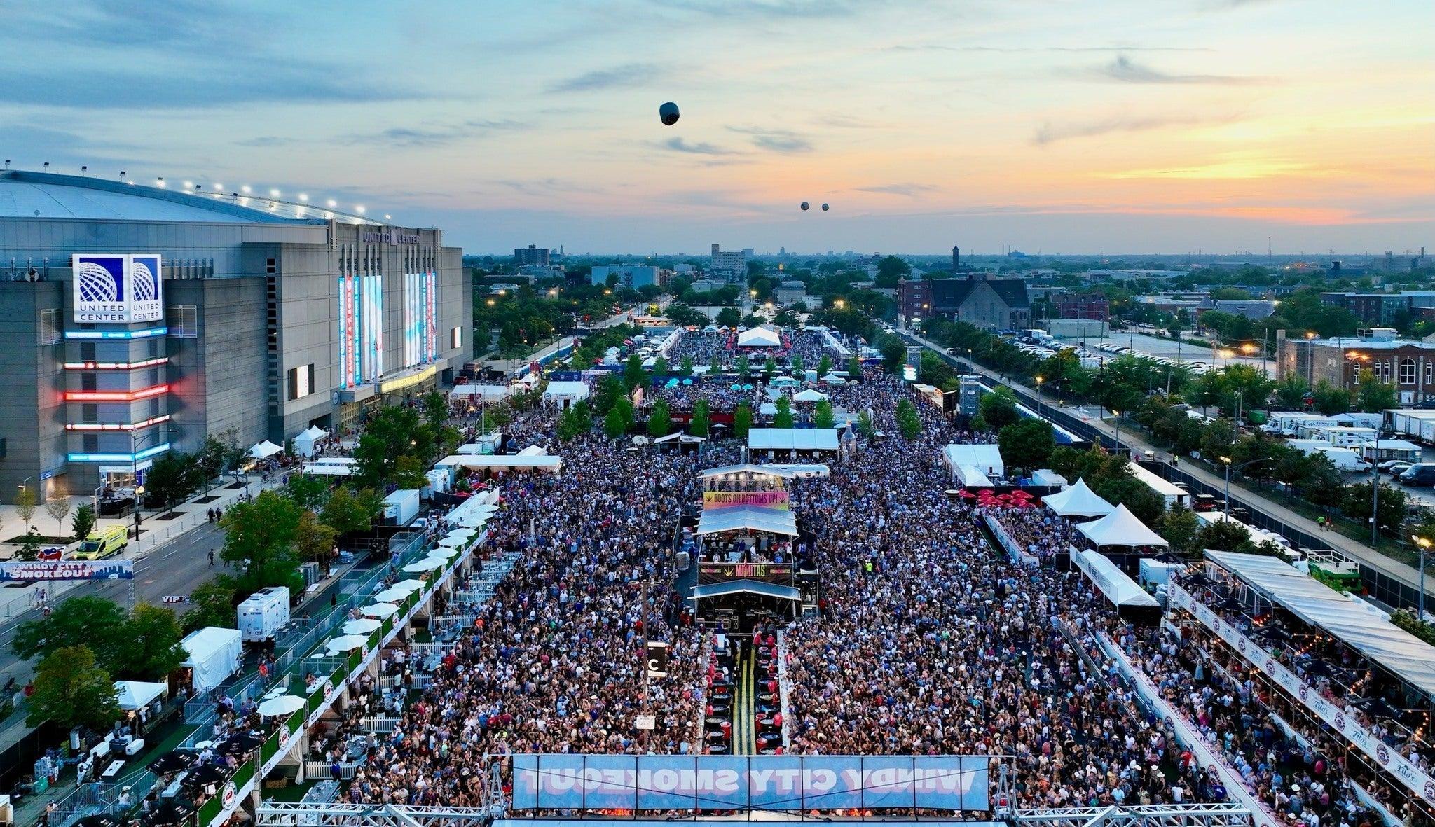 Aerial view of a massive outdoor country music festival crowd at dusk, with a stage, tents, and the United Center.