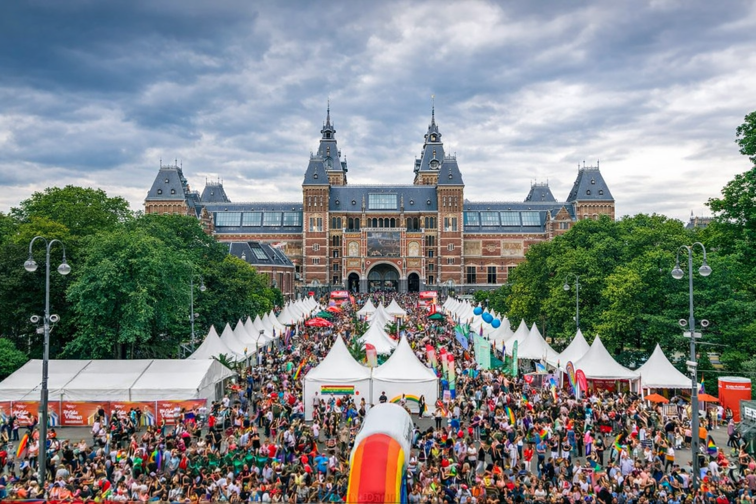 Crowd gathered for Pride in front of the Rijksmuseum, Amsterdam, with white tents and rainbow decorations.