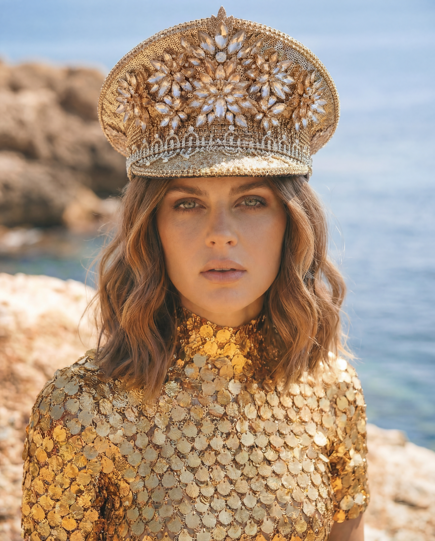 Glamorous woman in a shimmering gold sequin top and embellished captain's hat by the sea.