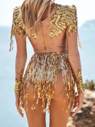 Gold rhinestone sequin tassel bodysuit with sheer sleeves and back, worn by a woman facing away.