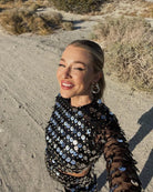 Vrouw draagt een zwarte mini-feestjurk met pailletten en uitsparingen, glimlacht buiten terwijl de pailletten schitteren in het zonlicht.