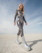 Woman in a black sequin jumpsuit with silver rhinestones and platform boots, walking in a desert.