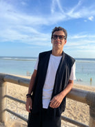 Mann in einem ärmellosen schwarzen Cardigan am Strand, der einen entspannten Boho-Stil zeigt.