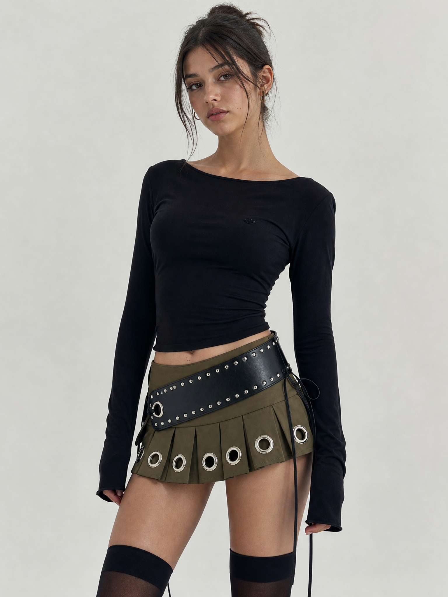 Model in a black long-sleeve top and an olive green mini skirt with large silver grommets and a studded black belt.