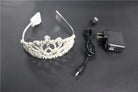 Silver rhinestone LED tiara with a flexible band, accompanied by a black USB charging cable and power adapter.