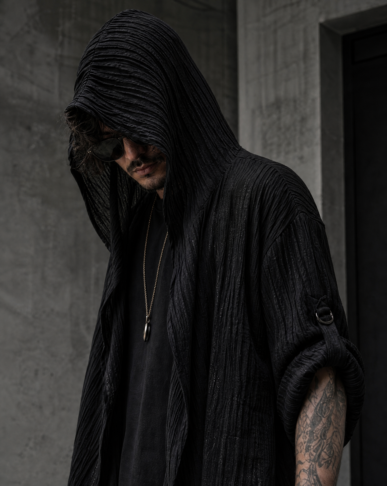 Man wearing a black, textured, long-sleeved hooded cardigan with a draped design and rolled sleeves in a modern urban setting.