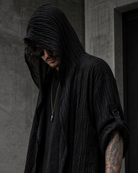 Man wearing a black, textured, long-sleeved hooded cardigan with a draped design and rolled sleeves in a modern urban setting.