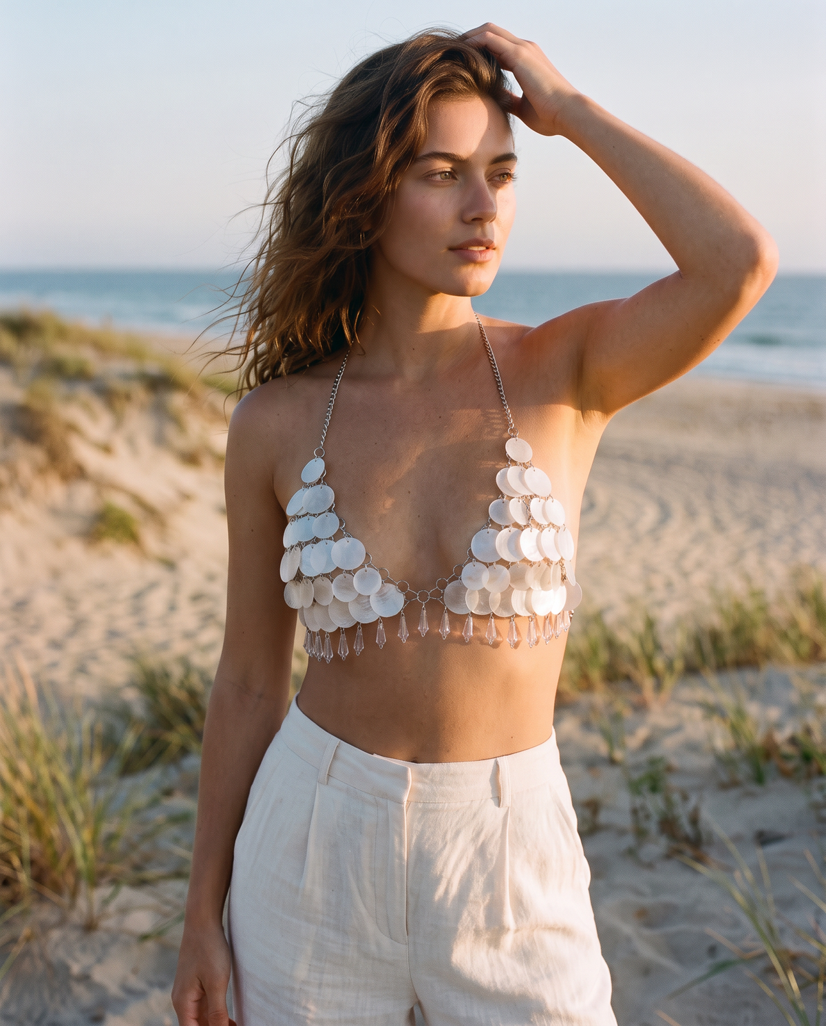 White seashell bra top featuring layered iridescent shells and delicate silver metal chains on a beach background.